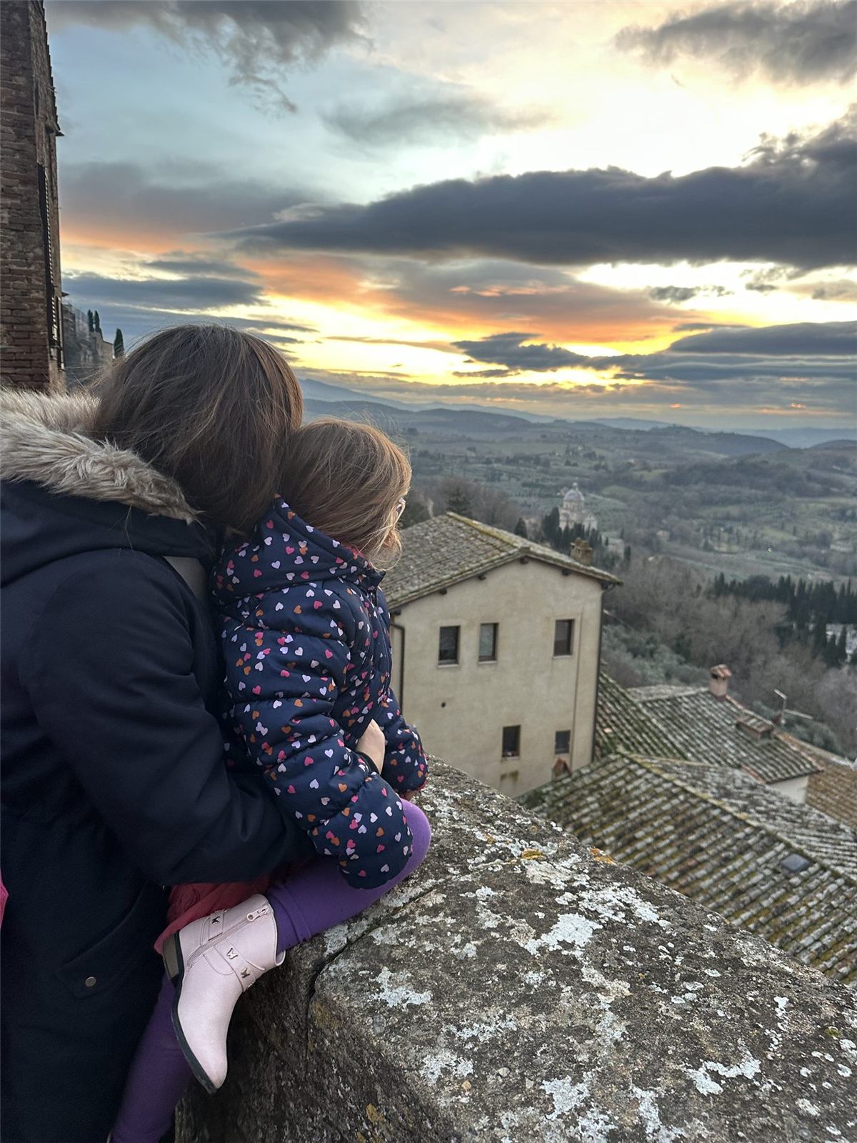 Kailah holding one of the girls on a stone wall, looking out over terracotta roofs and Tuscan hills at sunset near Pistoia