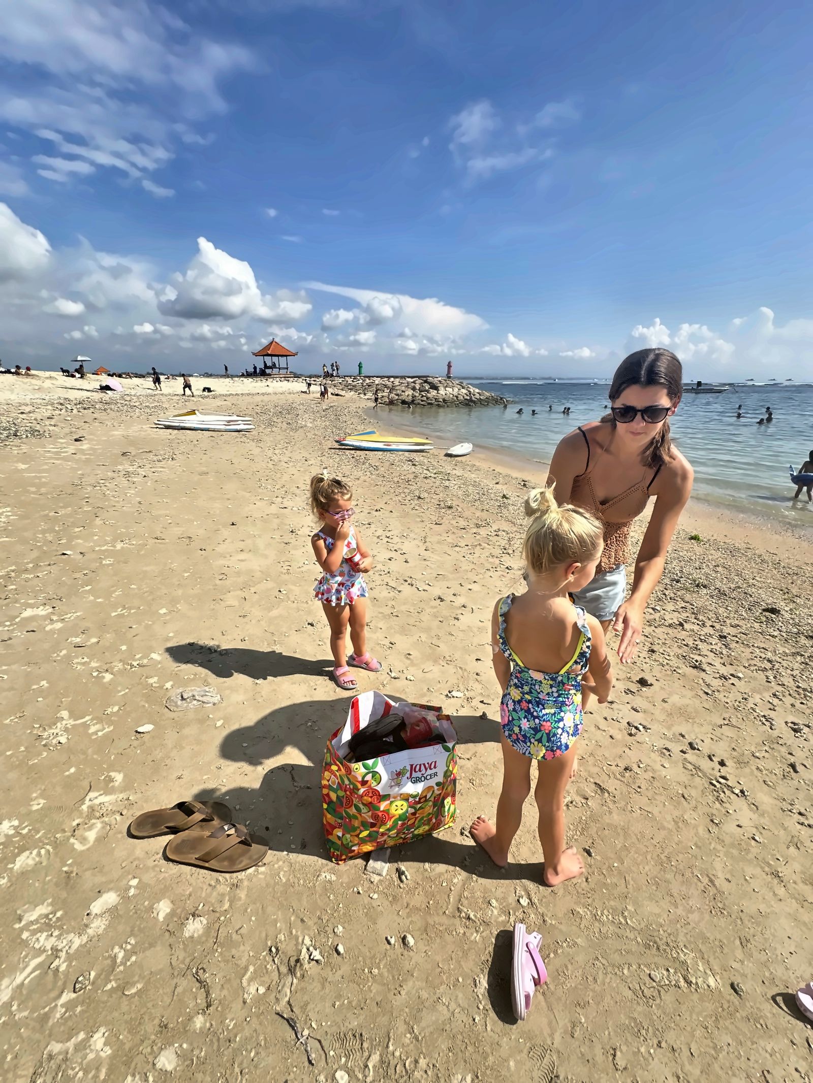 Kailah with the two girls in swimsuits on Sanur Beach in Bali, with outrigger boats and a thatched pavilion behind them